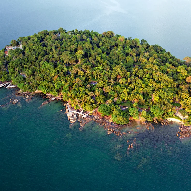 An island with vibrant greenery and dense trees, surrounded by tranquil blue ocean and natural rocky shoreline, seen from an overhead perspective.