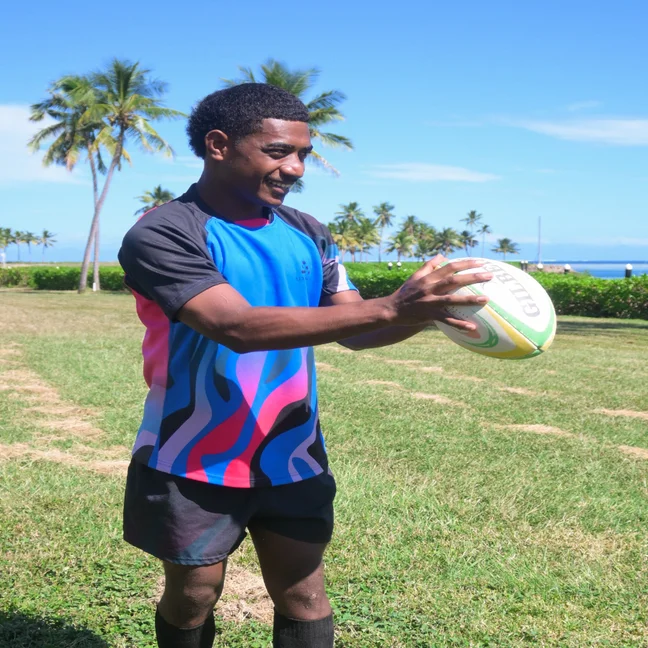 Person in a vibrant rugby uniform stands on a green field, holding a rugby ball. Palm trees and clear blue sky create a natural outdoor backdrop.