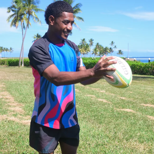 Person in a vibrant rugby uniform stands on a green field, holding a rugby ball. Palm trees and clear blue sky create a natural outdoor backdrop.