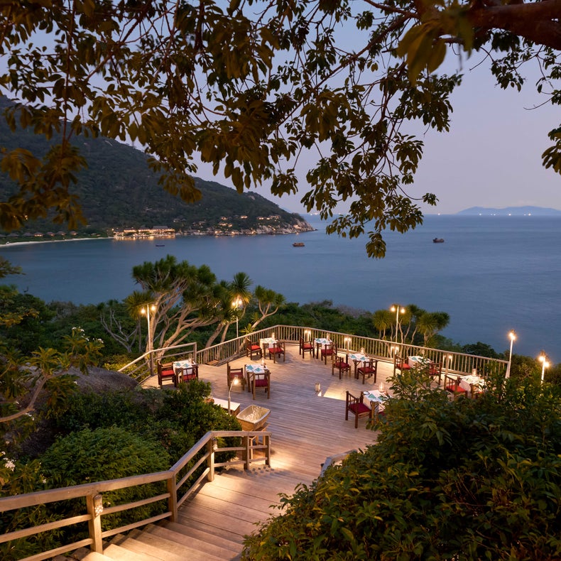 Salle à manger avec tables et chaises sur une terrasse bordée de verdure et de collines lointaines. Un éclairage d'ambiance permet de se détendre à la tombée de la nuit.