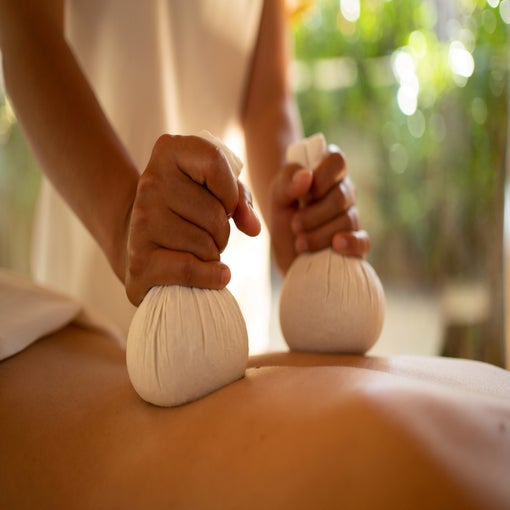 Herbal compress balls are gently applied to a guest’s back during a wellness massage, with soft daylight and garden views connecting the space to nature.