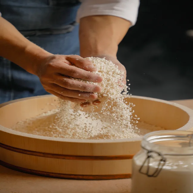 Hands gently sifting rice over a wooden bowl, grains cascading down and a glass jar set to the side, illustrating mindful preparation and care.