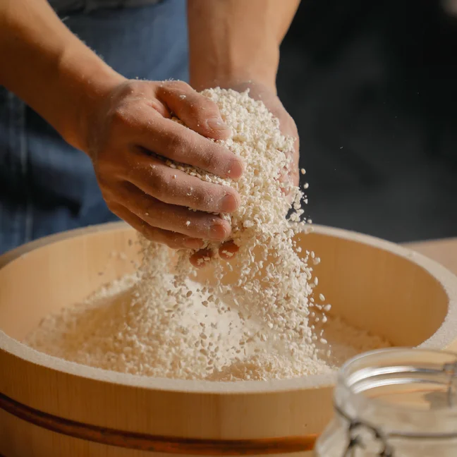 Hands gently sifting rice over a wooden bowl, grains cascading down and a glass jar set to the side, illustrating mindful preparation and care.