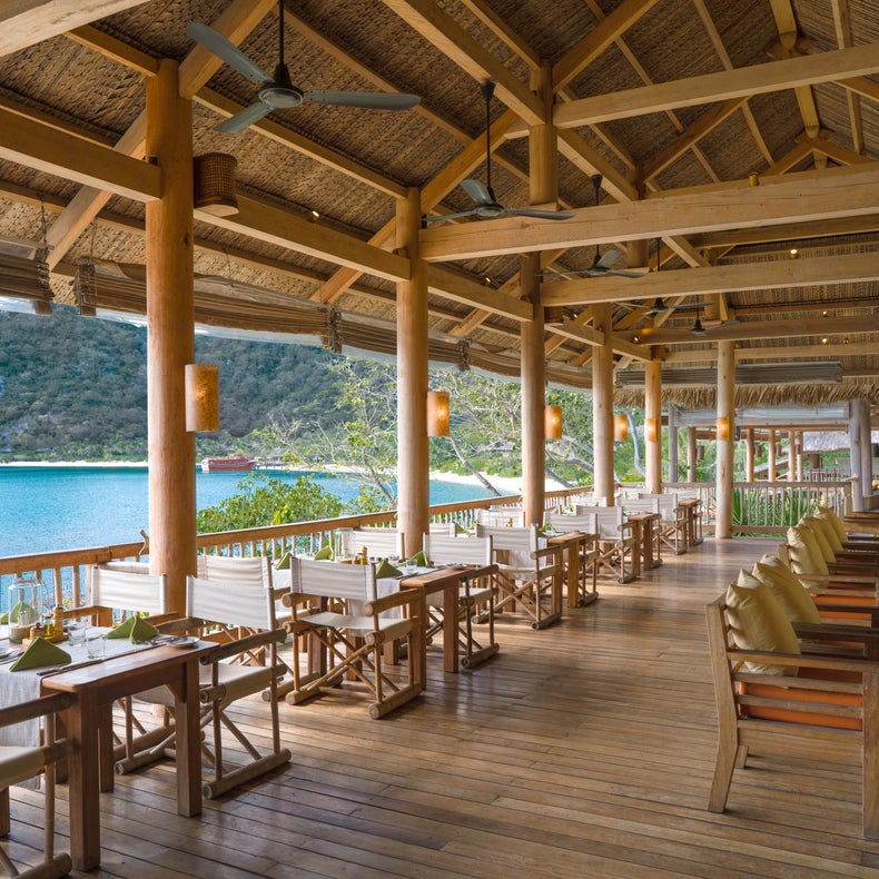 Restaurant avec éléments en bois naturel, grandes tables et chaises, donnant sur un lac paisible et les collines boisées en arrière-plan.