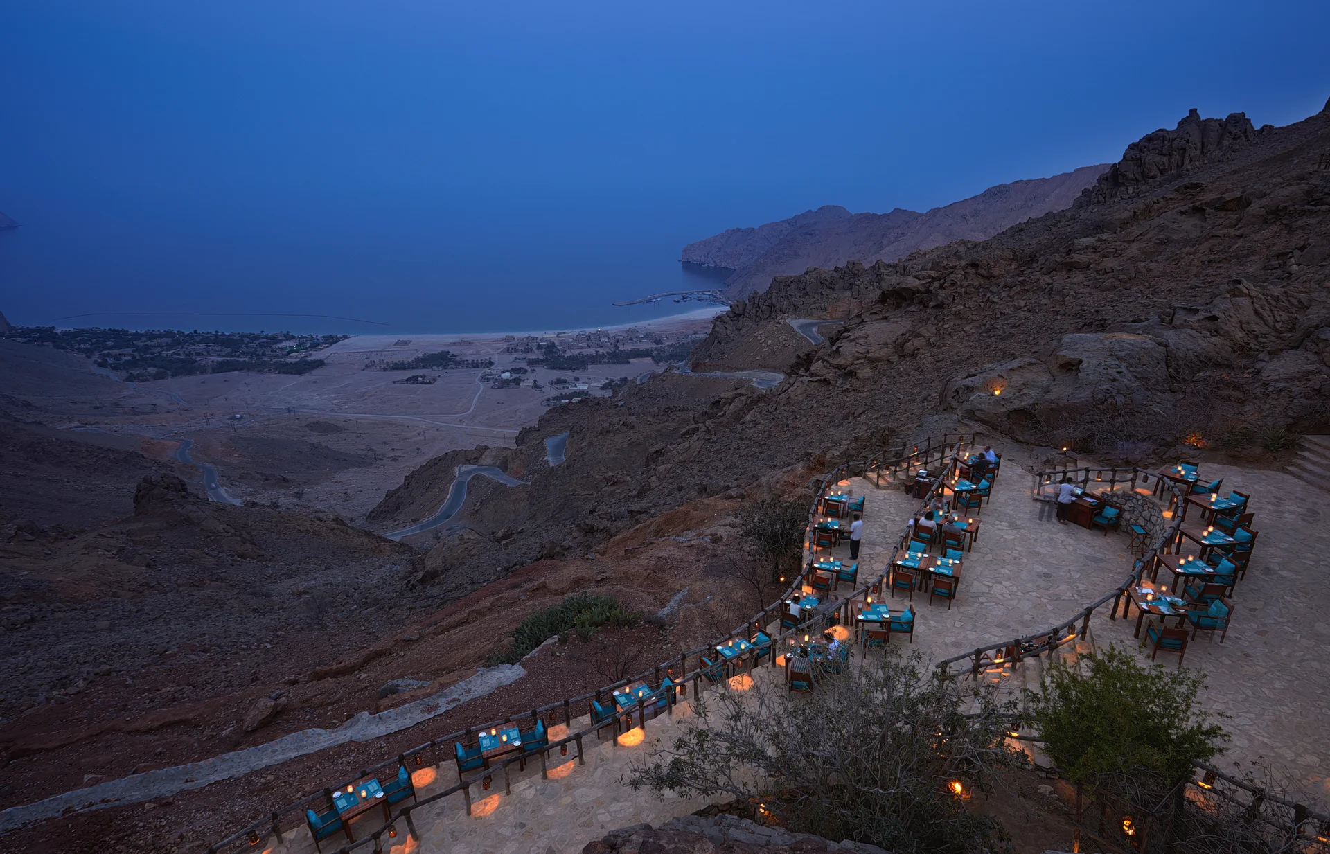 Sense on the Edge at dusk, candlelit tables strung along a narrow railed clifftop terrace high above the bay, with the resort and its beach visible far below on the plain.
