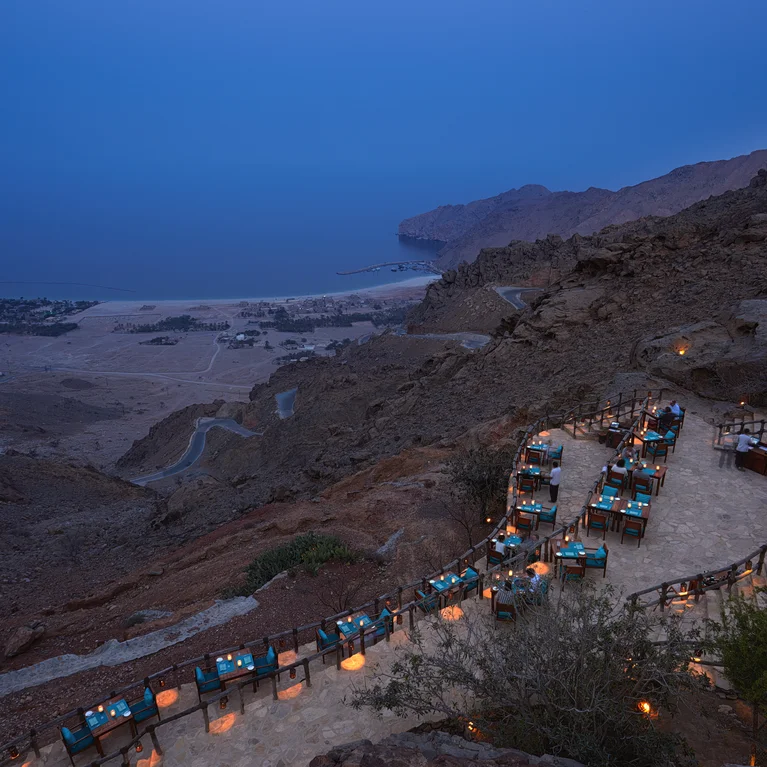 Mesas de comedor con manteles azules bordean un sendero de montaña al atardecer, ofreciendo vistas del mar, las verdes colinas y el pueblo en la distancia.