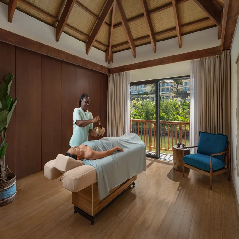 Guest enjoys a wellness treatment on a massage table in a calming room with large windows, natural wood floors, and views of lush, green gardens.