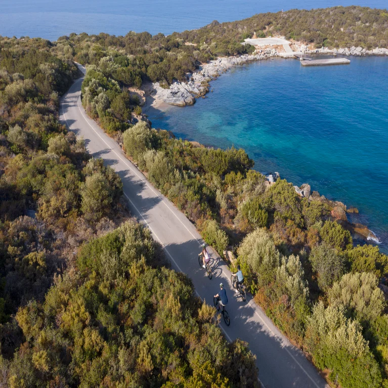 Three cyclists ride along a coastal road bordered by lush greenery, with the blue sea and rocky shoreline visible nearby.