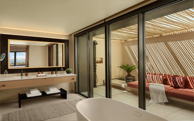 Bathroom featuring dual sinks, spacious mirror, soaking tub, and glass doors leading to a patio with red cushion seating, greenery, and wooden pergola.