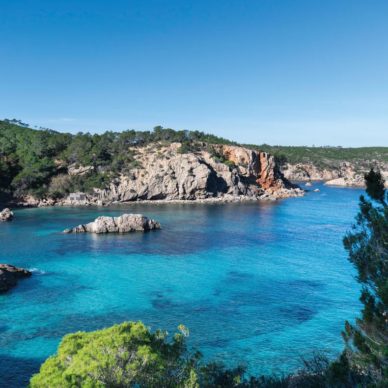 Xarraca Bay from the cliff above, turquoise water over pale rock shelves with a small islet offshore and pine-covered headlands running out to sea.