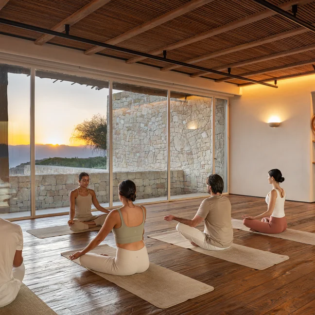 Guests participate in a group meditation or yoga session on mats in a serene room with stone walls and large windows framing the sunset outside.