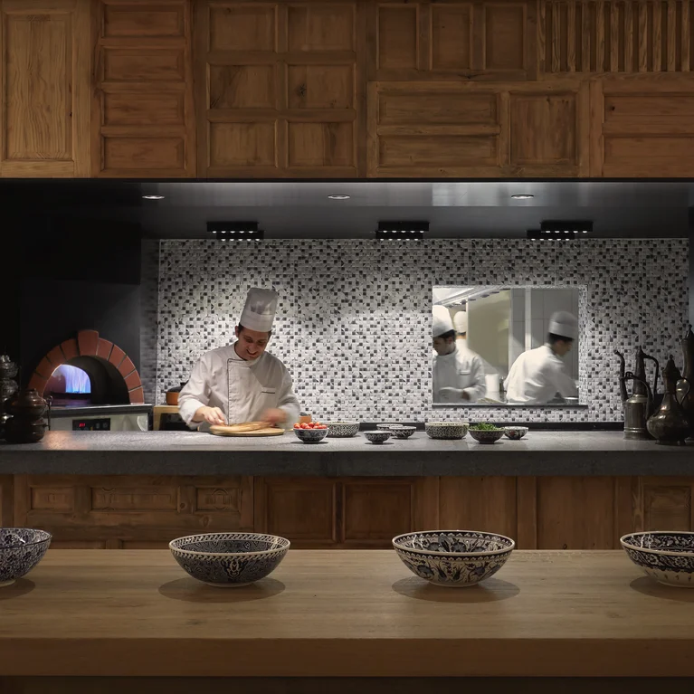Un chef vestido de blanco detrás de un moderno mostrador de cocina decorado con azulejos de mosaico; dos miembros del equipo trabajando en la parte trasera, cuencos decorados sobre un mostrador de madera en la parte delantera.