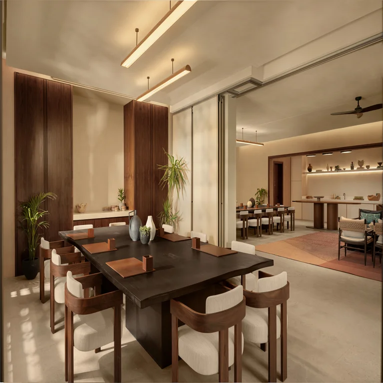 Dining space with a dark wood table, white cushioned chairs, greenery accents, and gentle lighting. Sliding doors open to a second dining area beyond.