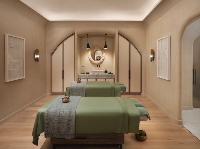 Spa room with two massage tables draped in green sheets, gentle lighting, round mirror, and simple modern wall art fostering a restorative atmosphere.