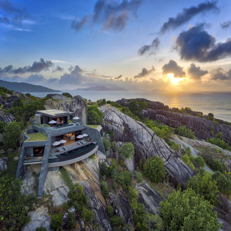 Eco-conscious residence set atop natural cliffs, framed by lush foliage, with expansive ocean views beneath a sky painted by the soft light of sunset.