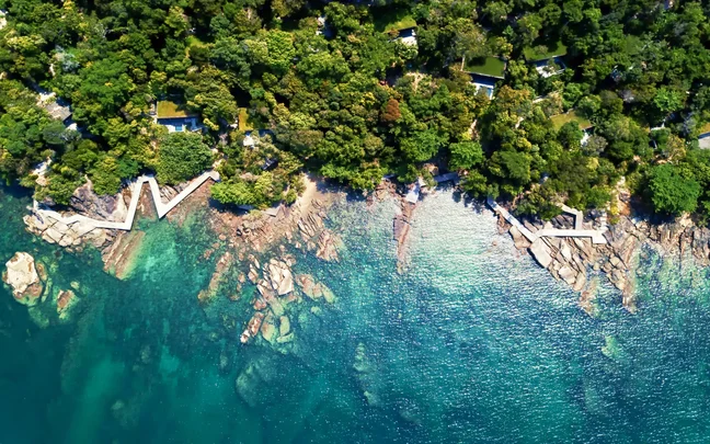 Green forest meets clear turquoise water along a rocky shoreline, with discreet white buildings thoughtfully positioned among the trees.