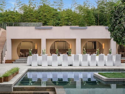 モダンな建物の横に白いダイニングテーブルと椅子が置かれ、青々とした植物に囲まれた穏やかなプールに映えている。.