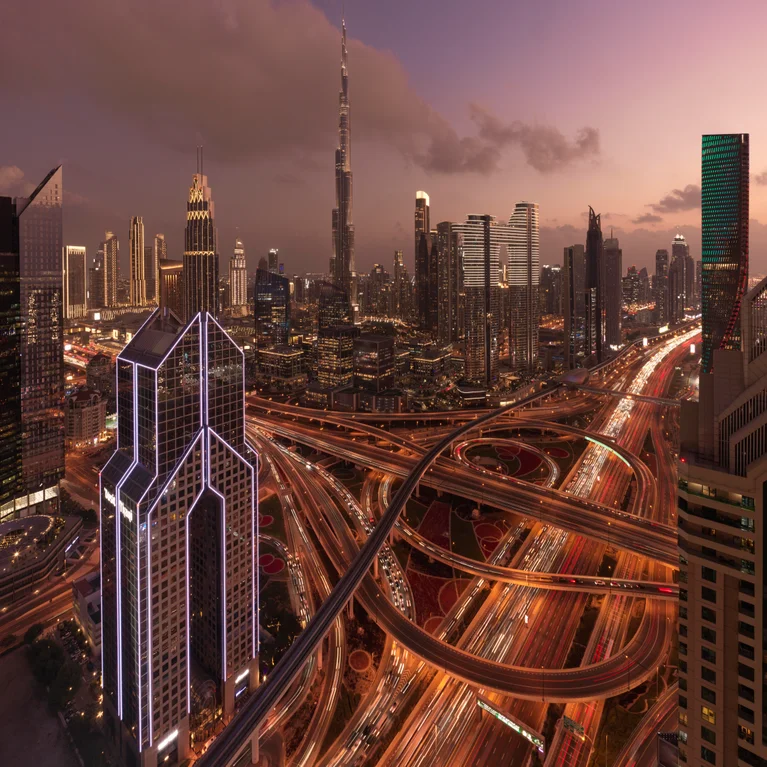 Dubai city skyline at dusk with the Burj Khalifa, surrounding skyscrapers, and softly lit highways showing gentle lines of moving traffic.
