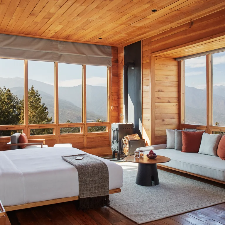Contemporary wooden bedroom with large windows, bed, and sofa. Mountain views visible outside. Natural light enhances neutral, minimalist furnishings and inviting atmosphere.