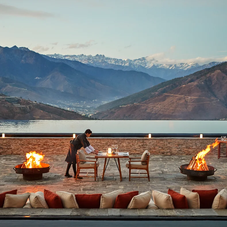 A set dining table sits by fire pits on a stone patio, with mountains and an infinity pool visible at sunset. Cushioned seating is arranged in the foreground.