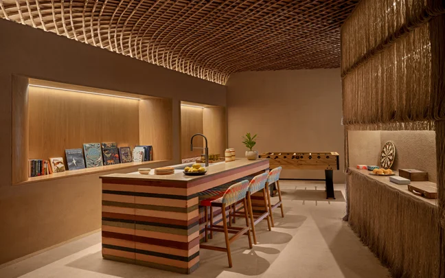 A communal lounge space featuring a kitchen island with stools, a foosball table, floor-to-ceiling bookshelves, and a slatted wood ceiling.