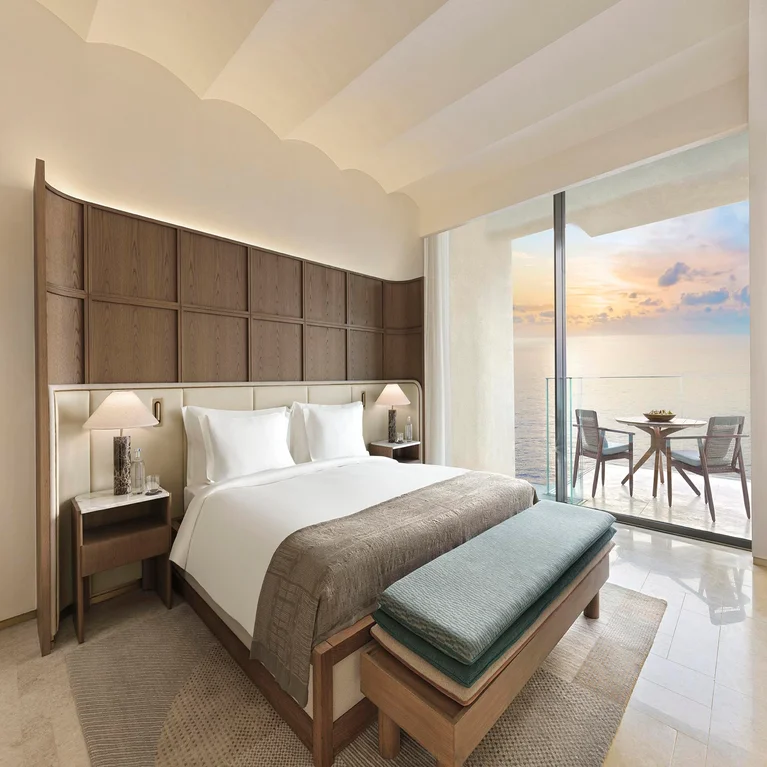 Spacious room featuring natural wood details, a generous bed, a bench, and balcony seating with views of sunlit ocean waters. Accessible pathways throughout the space.