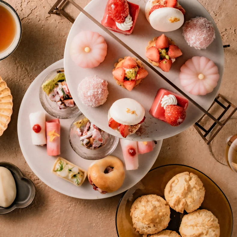 マカロン、スコーン、餅など、芸術的に盛り付けられたデザートが、陶器の皿と急須に盛られたお茶とともに、ナチュラルな色調のテーブルの上に供される。.