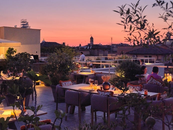 Open-air dining area with comfortable seating, abundant greenery, softly illuminated tables, and guests enjoying views of city rooftops beneath a vibrant evening sky.