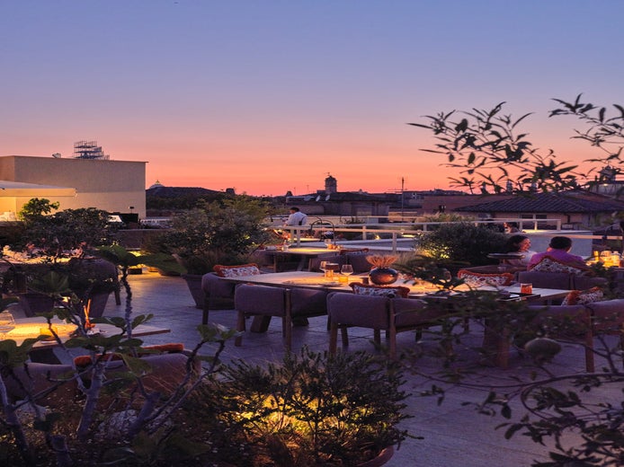 Open-air dining area with comfortable seating, abundant greenery, softly illuminated tables, and guests enjoying views of city rooftops beneath a vibrant evening sky.