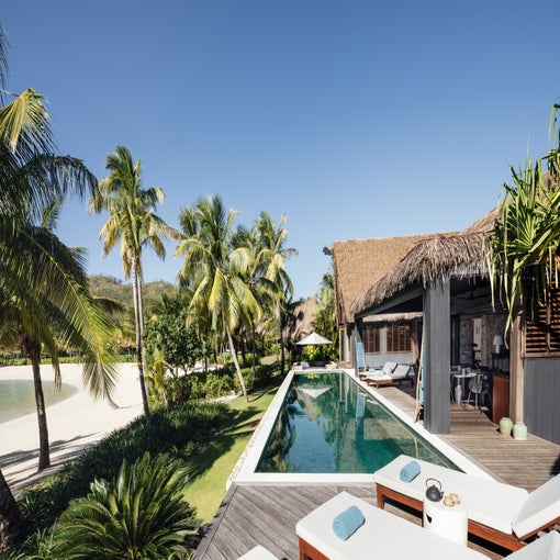Private villa featuring a pool, shaded by palm trees, with sun loungers and direct access to the beach. Clear sky enhances natural light and serene atmosphere.