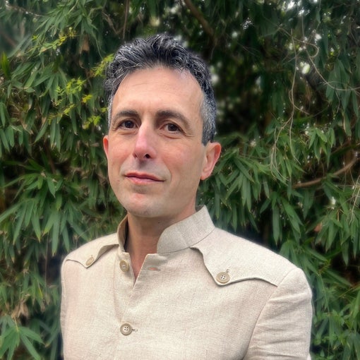 Individual with short, dark hair in a beige shirt stands before vibrant green foliage, gazing ahead. Neutral colors and natural setting reflect calm and simplicity.