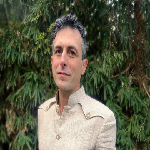Individual with short, dark hair in a beige shirt stands before vibrant green foliage, gazing ahead. Neutral colors and natural setting reflect calm and simplicity.