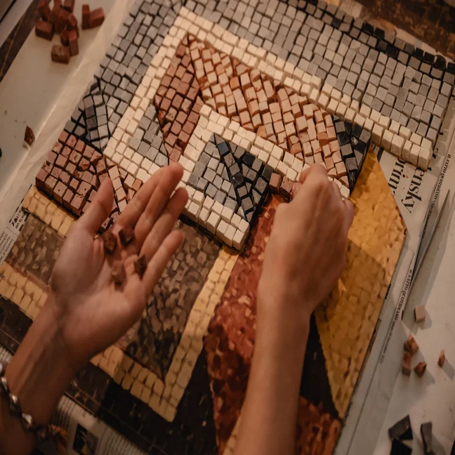 Hands mindful arrange red, yellow, cream, and gray mosaic tiles across a geometric pattern, reflecting thoughtful attention to detail in artful creation.