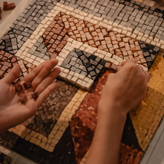Hands mindful arrange red, yellow, cream, and gray mosaic tiles across a geometric pattern, reflecting thoughtful attention to detail in artful creation.